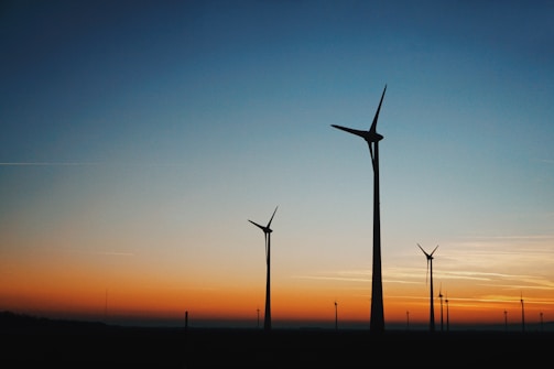 Silhouettes of wind turbines stand against a vibrant sunset sky, transitioning from a deep orange near the horizon to a clear blue as it rises.