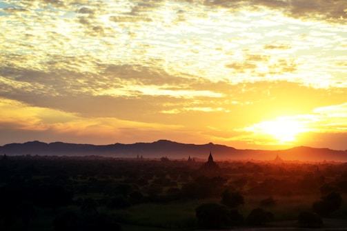 The image captures a stunning sunset over a landscape dotted with ancient temples. The sky is filled with dramatic clouds, illuminated in shades of orange and yellow as the sun descends towards the horizon. The foreground shows a vast landscape with scattered greenery and prominent silhouettes of pointed temple structures.
