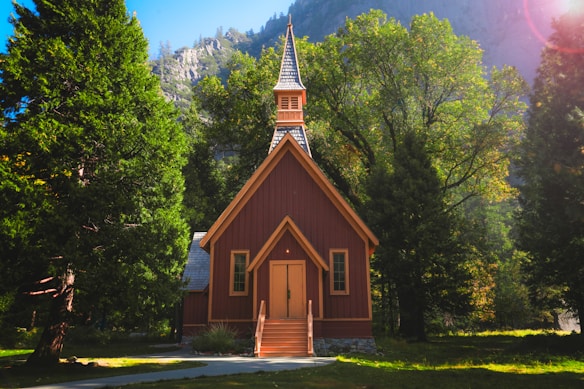 A small, picturesque church with a steep gabled roof and a pointed steeple, nestled among lush green trees. Sunlight filters through the leaves, casting gentle shadows on the church's wooden structure. The backdrop features a rocky mountain, enhancing the serene and secluded atmosphere.