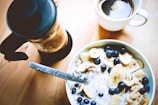 A delicious breakfast setting includes a bowl of cereal topped with banana slices, blueberries, and nuts. Next to it sit a cup of coffee and a French press on a wooden table. The morning light softly illuminates the scene, giving it a cozy and inviting feel.