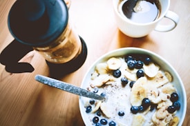 A delicious breakfast setting includes a bowl of cereal topped with banana slices, blueberries, and nuts. Next to it sit a cup of coffee and a French press on a wooden table. The morning light softly illuminates the scene, giving it a cozy and inviting feel.