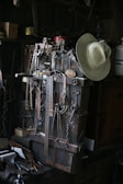 A collection of vintage metal tools is arranged on a workbench, including various hooks, wrenches, and measuring devices. A weathered cowboy hat is hung over the tools, adding a rustic touch. The setting is dimly lit, casting shadows over the well-used items, showcasing an atmosphere of history and craftsmanship.