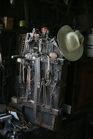 A collection of vintage metal tools is arranged on a workbench, including various hooks, wrenches, and measuring devices. A weathered cowboy hat is hung over the tools, adding a rustic touch. The setting is dimly lit, casting shadows over the well-used items, showcasing an atmosphere of history and craftsmanship.