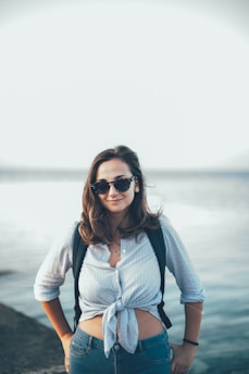 A person wearing sunglasses and a striped shirt tied at the waist stands confidently with hands on hips. The background features a calm, expansive body of water under a clear sky.