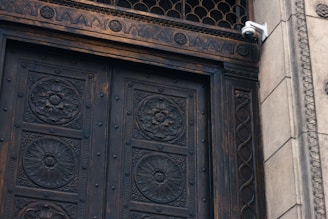 A detailed close-up of an ornate wooden door featuring intricate floral carvings and metal rivets. The door is framed by decorative stonework with architectural patterns. A white security camera is mounted on the upper right corner of the stone frame, adding a modern element to the historical design.