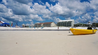 A sandy beach with a blue sky dotted with fluffy clouds. Several people are enjoying the area under beach umbrellas and chairs. A bright yellow kayak lies on the sand close to the water. Modern beachfront buildings line the background.