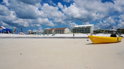 A sandy beach with a blue sky dotted with fluffy clouds. Several people are enjoying the area under beach umbrellas and chairs. A bright yellow kayak lies on the sand close to the water. Modern beachfront buildings line the background.