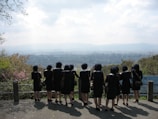 A group of people wearing dark uniforms and hats stand in a row, facing away from the camera. They overlook a vast cityscape with mountains in the distance. The sky is partly cloudy, and the area is surrounded by greenery.