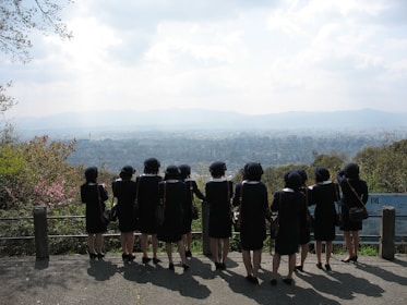 A group of people wearing dark uniforms and hats stand in a row, facing away from the camera. They overlook a vast cityscape with mountains in the distance. The sky is partly cloudy, and the area is surrounded by greenery.