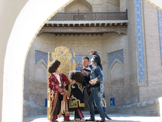 A group of people is gathered around a decorative, ornate chair placed against a backdrop of traditional, geometrically patterned brick walls. The individuals are dressed in colorful, elaborate costumes, including large, curly wigs and embroidered garments, indicating a cultural or historical theme.
