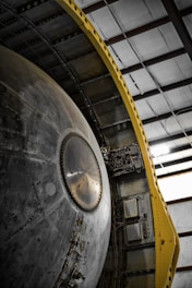 A close-up view of a large mechanical structure with metallic surfaces, featuring intricate details and components. There is a prominent circular element in the center with a series of rivets around it. The structure is flanked by a yellow support frame on one side, contrasting with the metal. The setting is an industrial or warehouse environment with visible steel beams above.