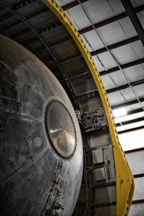 A close-up view of a large mechanical structure with metallic surfaces, featuring intricate details and components. There is a prominent circular element in the center with a series of rivets around it. The structure is flanked by a yellow support frame on one side, contrasting with the metal. The setting is an industrial or warehouse environment with visible steel beams above.