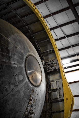 A close-up view of a large mechanical structure with metallic surfaces, featuring intricate details and components. There is a prominent circular element in the center with a series of rivets around it. The structure is flanked by a yellow support frame on one side, contrasting with the metal. The setting is an industrial or warehouse environment with visible steel beams above.