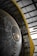 A close-up view of a large mechanical structure with metallic surfaces, featuring intricate details and components. There is a prominent circular element in the center with a series of rivets around it. The structure is flanked by a yellow support frame on one side, contrasting with the metal. The setting is an industrial or warehouse environment with visible steel beams above.