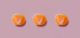 Three orange citrus fruit halves are evenly spaced in a row against a muted pink background. The fruits have a bright, juicy appearance with a contrast between the vibrant orange of the segments and the subtle pink backdrop.