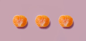Three orange citrus fruit halves are evenly spaced in a row against a muted pink background. The fruits have a bright, juicy appearance with a contrast between the vibrant orange of the segments and the subtle pink backdrop.