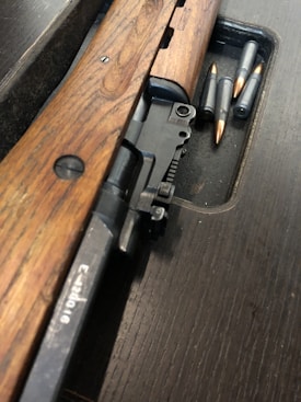 A wooden rifle with metallic components is placed on a dark wooden surface, accompanied by several bullets. The rifle showcases intricate metal parts with a visible bolt action mechanism.
