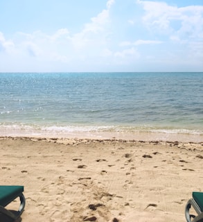 A serene beach scene with light beige sand and two green sunbeds partially visible on both sides. The horizon features a calm blue sea that meets a slightly clouded sky with varying shades of blue and white.