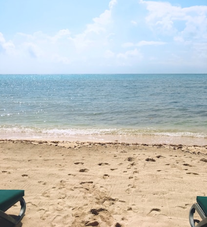 A serene beach scene with light beige sand and two green sunbeds partially visible on both sides. The horizon features a calm blue sea that meets a slightly clouded sky with varying shades of blue and white.