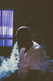 A person is sitting indoors, wearing a white shirt. They are exhaling a large amount of smoke, which is illuminated by a light source. The setting appears dimly lit, with an open window in the background revealing blue curtains.