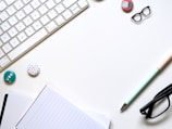 A minimalist workspace layout featuring a white keyboard, a lined notebook, and a pastel-colored pen. There are glasses, decorative badges, and a pair of patterned clips arranged neatly on the surface.