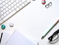 A minimalist workspace layout featuring a white keyboard, a lined notebook, and a pastel-colored pen. There are glasses, decorative badges, and a pair of patterned clips arranged neatly on the surface.