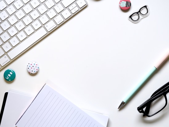 A minimalist workspace layout featuring a white keyboard, a lined notebook, and a pastel-colored pen. There are glasses, decorative badges, and a pair of patterned clips arranged neatly on the surface.