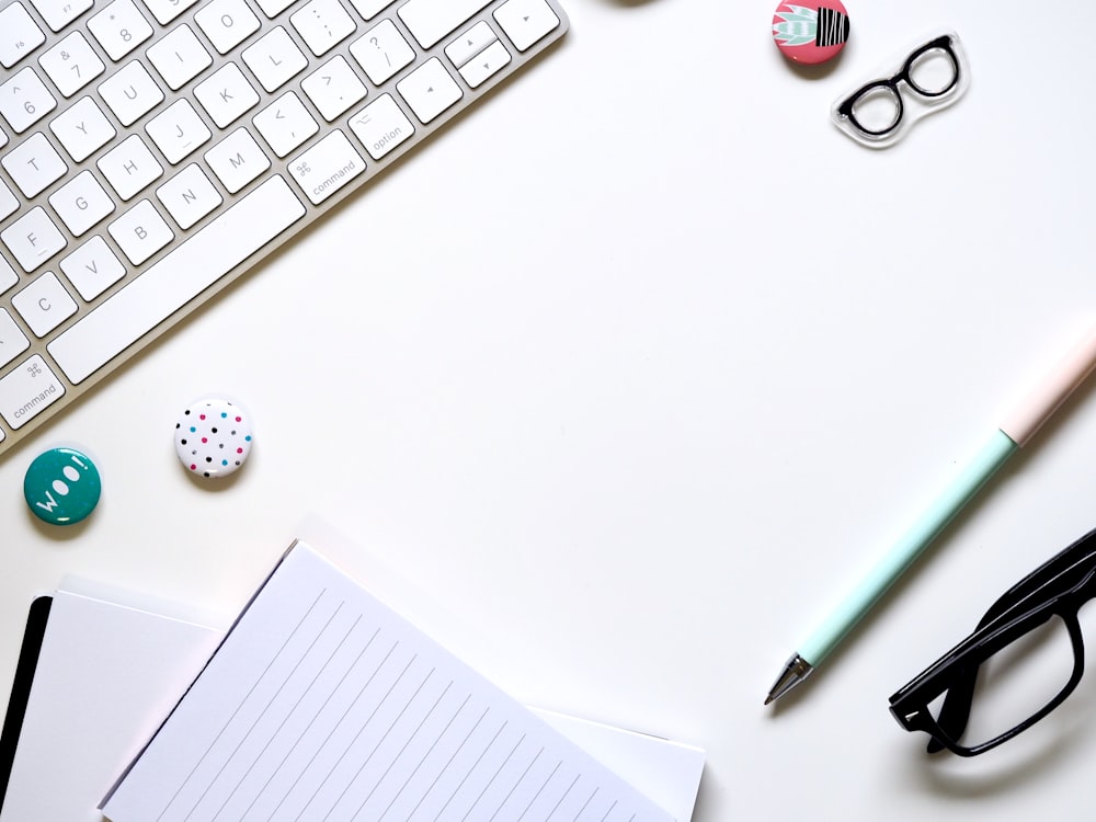 Du papier à lettres, un stylo bleu et des lunettes sur une surface blanche à côté du clavier
