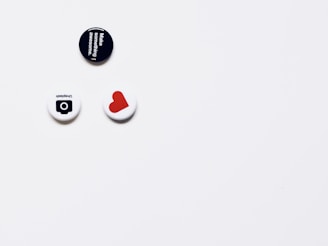 Three round pins or badges are placed on a white surface, each with distinct designs and colors. The top pin is black with white text that reads 'Make something awesome.' Below, two pins are aligned horizontally; one is white with a simple black design resembling a camera and the word 'Unsplash' upside down, and the other features a red heart shape on a white background.
