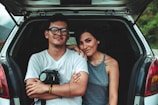 A young couple is sitting in the open trunk of a car. The man, wearing glasses and a white t-shirt, has a camera in his lap, while the woman, in a gray sleeveless top, leans affectionately against him. They are smiling calmly, appearing relaxed and content. The car is parked in a natural setting with a blurred view of greenery in the background.