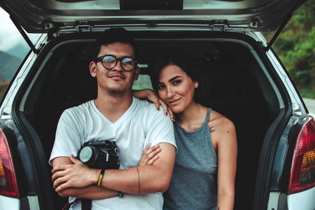 A young couple is sitting in the open trunk of a car. The man, wearing glasses and a white t-shirt, has a camera in his lap, while the woman, in a gray sleeveless top, leans affectionately against him. They are smiling calmly, appearing relaxed and content. The car is parked in a natural setting with a blurred view of greenery in the background.
