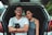A young couple is sitting in the open trunk of a car. The man, wearing glasses and a white t-shirt, has a camera in his lap, while the woman, in a gray sleeveless top, leans affectionately against him. They are smiling calmly, appearing relaxed and content. The car is parked in a natural setting with a blurred view of greenery in the background.