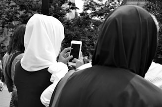 Two individuals in headscarves are viewed from behind, with one holding a smartphone in a public setting. Trees and other people are visible in the background.