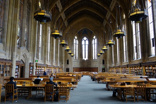 A grand library hall with tall arched windows and intricate chandeliers hanging from the vaulted ceiling. Long wooden tables surrounded by chairs fill the room, with people sitting and studying. Shelves of books line the walls, adding to the academic atmosphere.