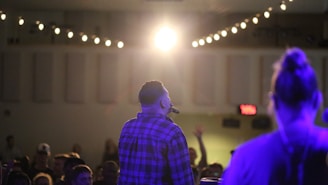 A man holding a microphone is facing a large audience in a dimly lit room. Overhead, string lights are illuminating the space, creating a warm atmosphere. Another person stands nearby, slightly out of focus.
