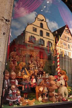 A display window featuring a collection of dolls, teddy bears, and toy animals dressed in traditional attire. Behind the toys, a backdrop depicts quaint European buildings, adding a nostalgic and whimsical atmosphere. Reflections of real buildings with architectural details are visible on the glass.