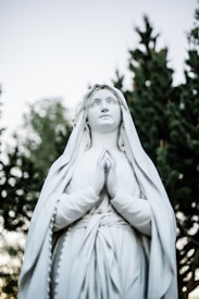 A statue of a serene figure with clasped hands in a gesture of prayer, wearing long robes and a veil. The statue is set against a backdrop of trees and a clear sky, suggesting an outdoor setting.