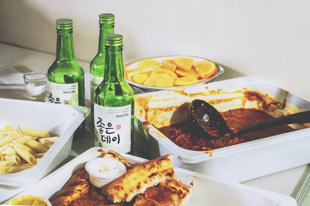 A variety of food items are arranged on a table, including a tray with a sauce and ladle, a plate of sliced lemons, a dish with uncooked penne pasta, and a plate with bread and a small container with white sauce. There are also four green glass bottles, likely containing a beverage, with Korean writing on their labels.
