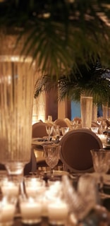 A beautifully decorated dining area with elegant table settings, featuring numerous candles and crystal glassware. Large pillars and lush green palm leaves contribute to a luxurious atmosphere.