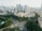 Aerial view of an urban landscape featuring a busy intersection surrounded by lush greenery and a cluster of high-rise buildings. Residential apartments with distinct yellow and beige tones are prominent in the foreground, while modern skyscrapers rise in the background against a hazy sky.