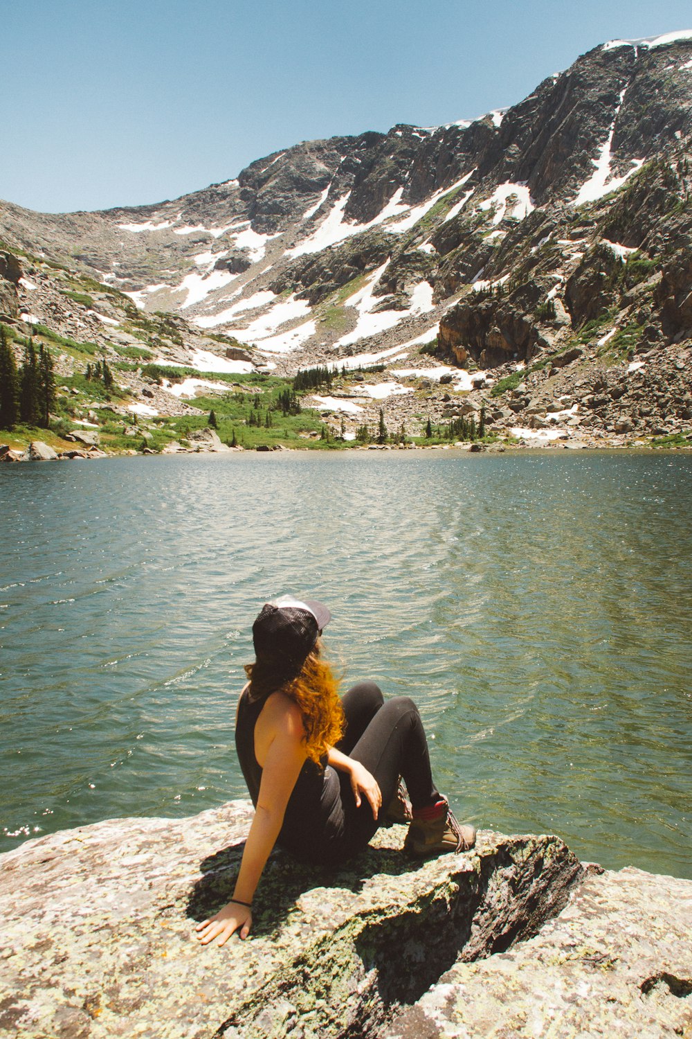 Mujer sentada en la formación rocosa mirando a través de la montaña nevada cerca del cuerpo de agua