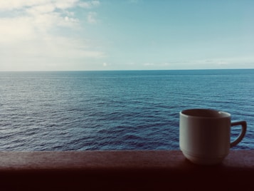 A serene view of the ocean with calm, blue water stretching to the horizon under a partly cloudy sky. In the foreground, a white ceramic mug sits on a wooden ledge, adding a sense of peaceful solitude.