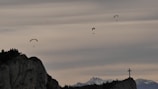 Paragliders are floating gracefully in the sky above a rocky landscape with mountains in the background. A large cross stands prominently on the right-hand side atop a cliff.