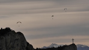 Paragliders are floating gracefully in the sky above a rocky landscape with mountains in the background. A large cross stands prominently on the right-hand side atop a cliff.