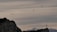 Paragliders are floating gracefully in the sky above a rocky landscape with mountains in the background. A large cross stands prominently on the right-hand side atop a cliff.