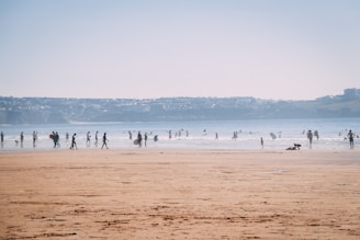 A sandy beach with numerous people walking, playing, and engaging in water activities near the shoreline. Surfboards are visible with surfers catching waves in the distance. The sky is clear, and the backdrop features distant hills or coastline.