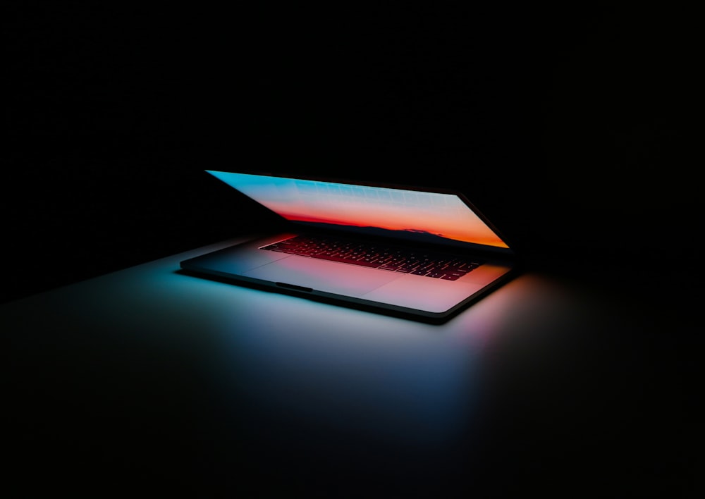 A refurbished Apple Macbook in a dark room with the screen half open illuminating the keybord in multiple colours