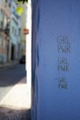 A street corner with a wall painted in light purple where the phrase 'GRL PWR' is written three times in green. The background shows a slightly blurred image of a narrow street with buildings and some greenery.