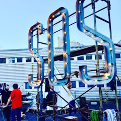 A large S.O.S. sign with light bulbs is mounted on a metal frame in front of a building with a corrugated metal exterior and windows. Two people stand nearby, one wearing a red shirt and the other with a face mask.