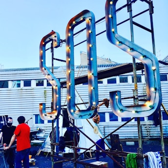 A large S.O.S. sign with light bulbs is mounted on a metal frame in front of a building with a corrugated metal exterior and windows. Two people stand nearby, one wearing a red shirt and the other with a face mask.